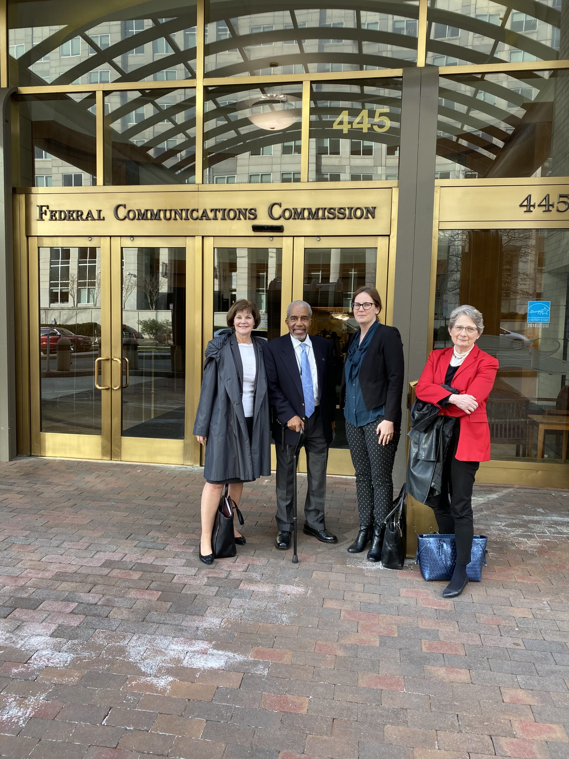 Standing in front of the FCC, from left to right, Loretta from WID, Robert Richardson, Rylin from AUCD, and Susan from PVA, posing and smiling at the camera